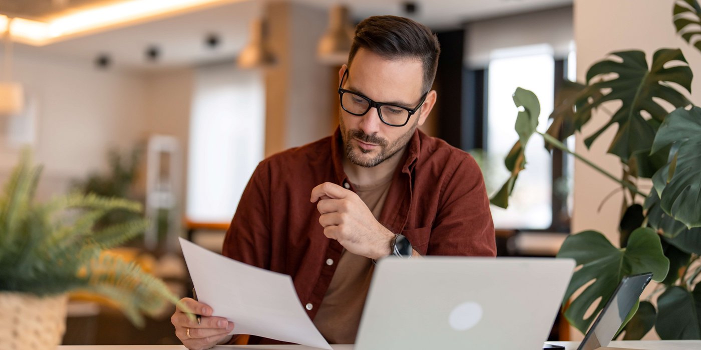 Ein Mann mit Brille arbeitet konzentriert am Schreibtisch. Er hält ein Dokument in der Hand, während ein Laptop vor ihm steht.