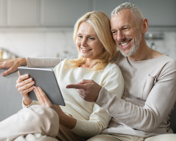 Ein älteres Paar sitzt entspannt auf dem Sofa und schaut gemeinsam lächelnd auf ein Tablet.