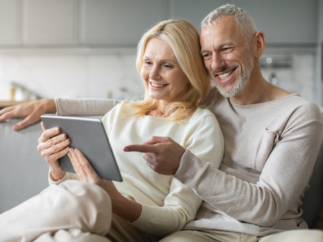 Ein älteres Paar sitzt entspannt auf dem Sofa und schaut gemeinsam lächelnd auf ein Tablet.