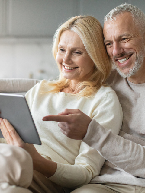 Ein älteres Paar sitzt entspannt auf dem Sofa und schaut gemeinsam lächelnd auf ein Tablet.