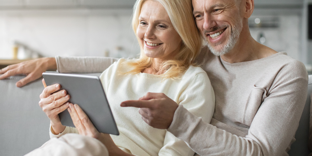 Ein älteres Paar sitzt entspannt auf dem Sofa und schaut gemeinsam lächelnd auf ein Tablet.
