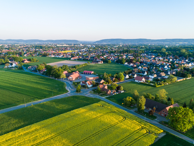 Luftaufnahme mit Wohnhäusern und umliegenden Feldern bei sonnigem Wetter.
