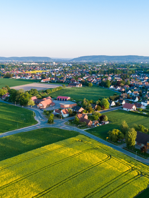Luftaufnahme mit Wohnhäusern und umliegenden Feldern bei sonnigem Wetter.