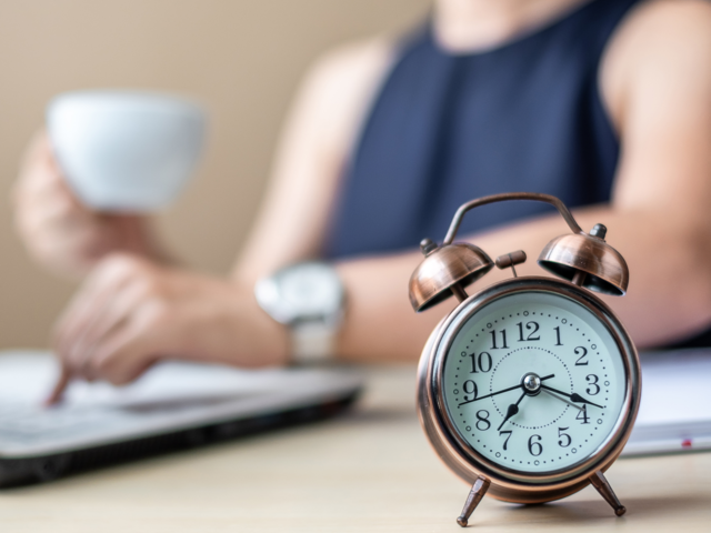 Frau im Hintergrund am Laptop mit Kaffeetasse in der Hand, im Vordergrund steht ein Wecker