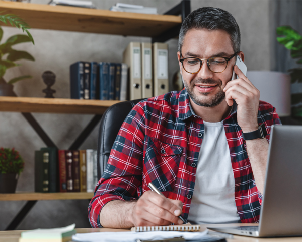 Mann sitzt am PC, telefoniert und macht sich Notizen