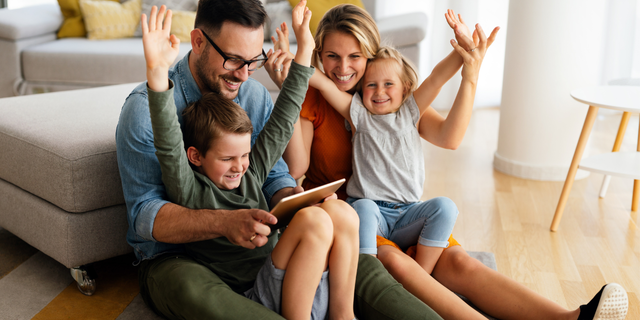 Familie sitzt gemeinsam im Wohnzimmer und nutzt ein Tablet, während Eltern und Kinder fröhlich die Hände heben.