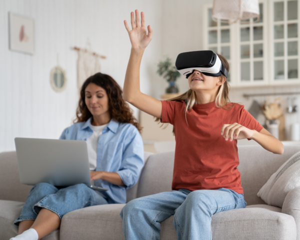 Mutter sitzt mit Laptop auf dem Schoß auf dem Sofa, daneben die Tochter mit einer VR Brille auf