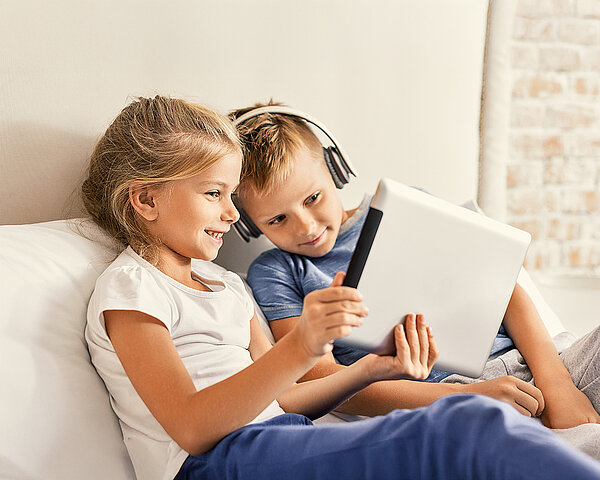 Zwei Kinder sitzen auf der Couch und schauen zusammen auf ein Tablet