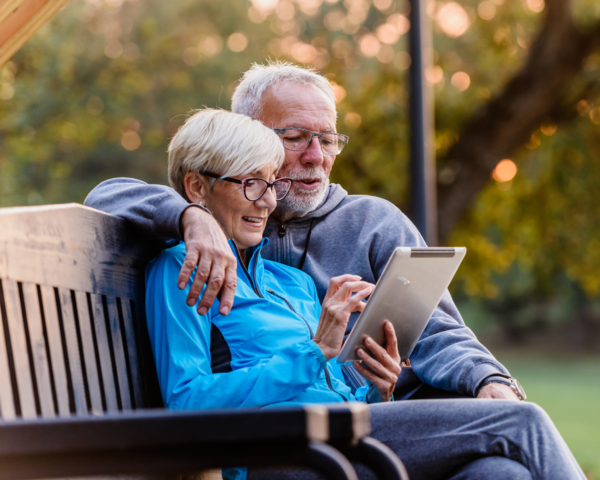 Ein älteres Paar sitzt draußen auf einer Bank und schaut gemeinsam auf ein Tablet.