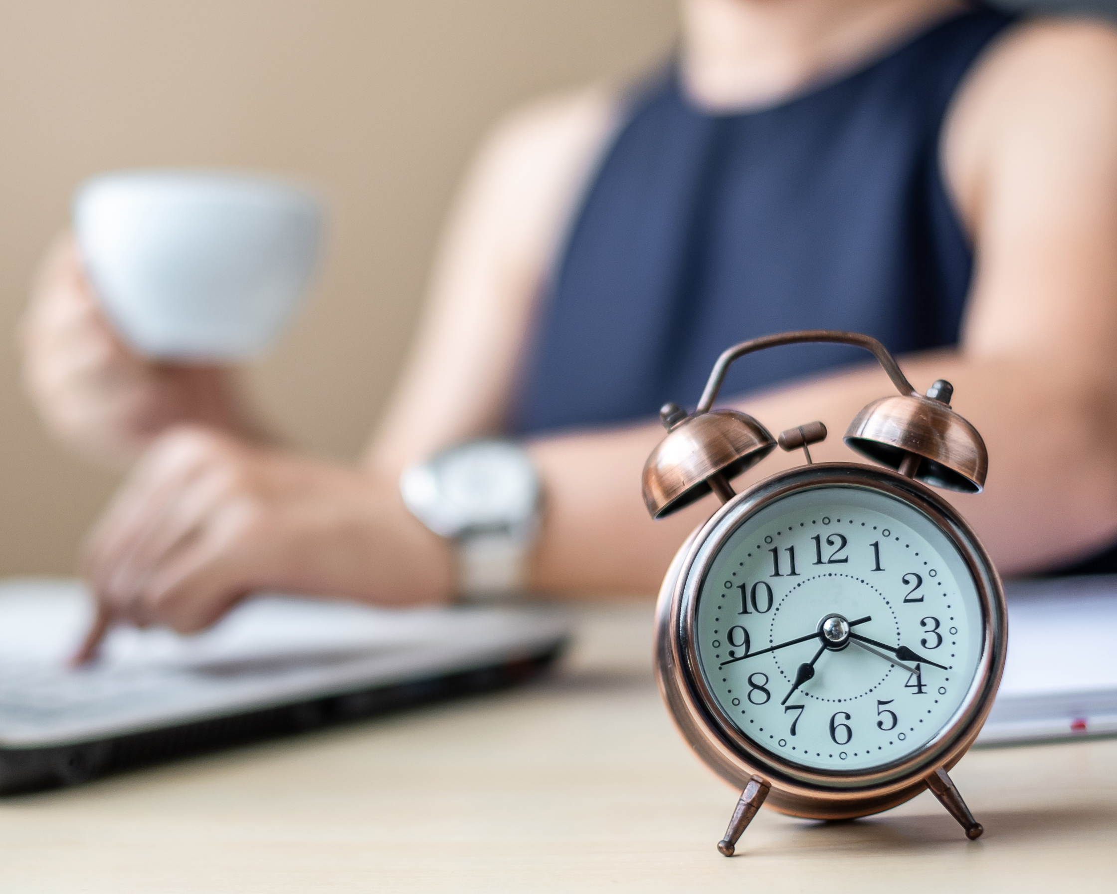 Frau im Hintergrund am Laptop mit Kaffeetasse in der Hand, im Vordergrund steht ein Wecker