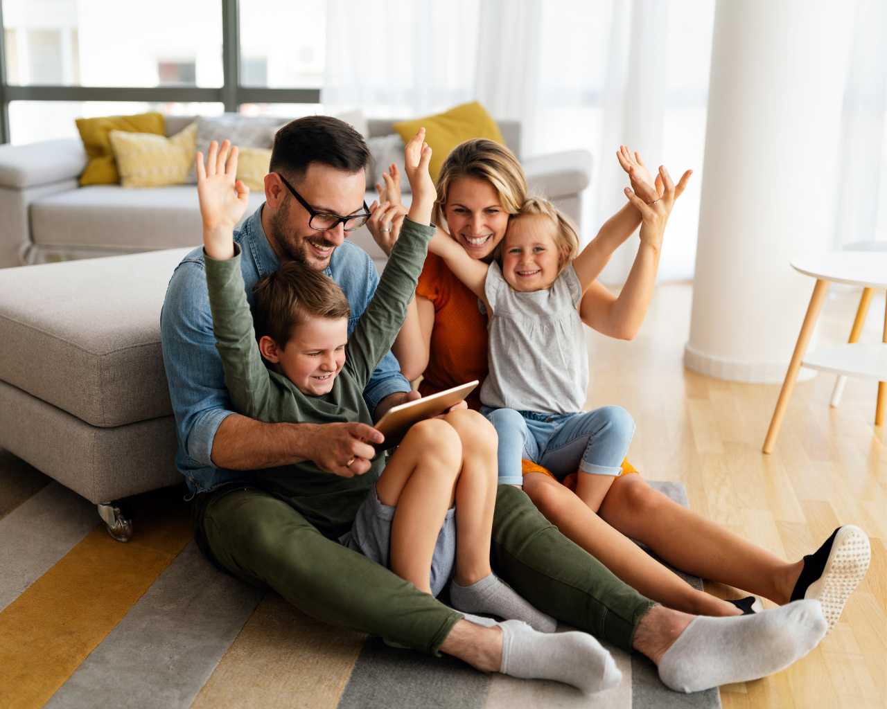 Familie sitzt gemeinsam im Wohnzimmer und nutzt ein Tablet, während Eltern und Kinder fröhlich die Hände heben.